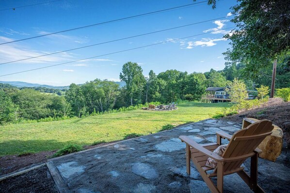 A patio with a stone table and wooden chairs, overlooking a grassy yard with a wooden deck and a house in the background.