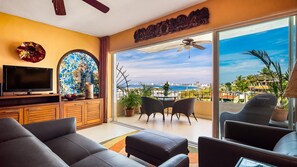 A living room with a TV, sofa, and chairs, a balcony with a view of the city and ocean, and a decorative wall. - Spectacular Condo w/ Ocean Views (Puerto Vallarta)