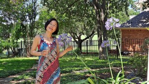 A woman in a patterned dress standing in a garden with a potted plant and a brick building in the background. - Comanche, Cameron,  Good Year, Fort-Sill, Medicine Park, Lawton, Oklahoma. 
 (Lawton)