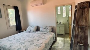 Een slaapkamer met een bed, airconditioning, plafondventilator, raam met gordijnen, een houten kledingkast en een zichtbare badkamer door een open deur.