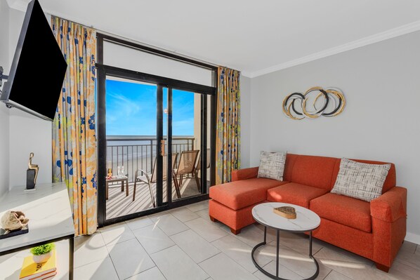 A modern living room with a red sofa, a round coffee table, a flat-screen TV, and a balcony with a view of the sea.