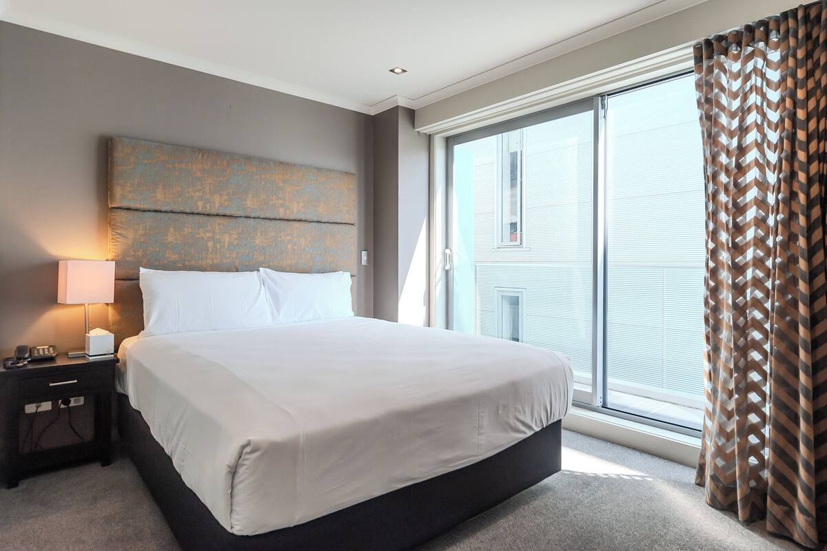 A hotel room with a large bed, a nightstand with a lamp, and a view of the outside through a sliding door.