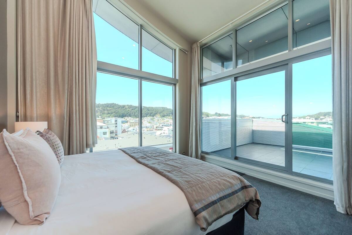 A hotel room with a large bed, a view of the city through floor-to-ceiling windows, and a balcony with a green roof.
