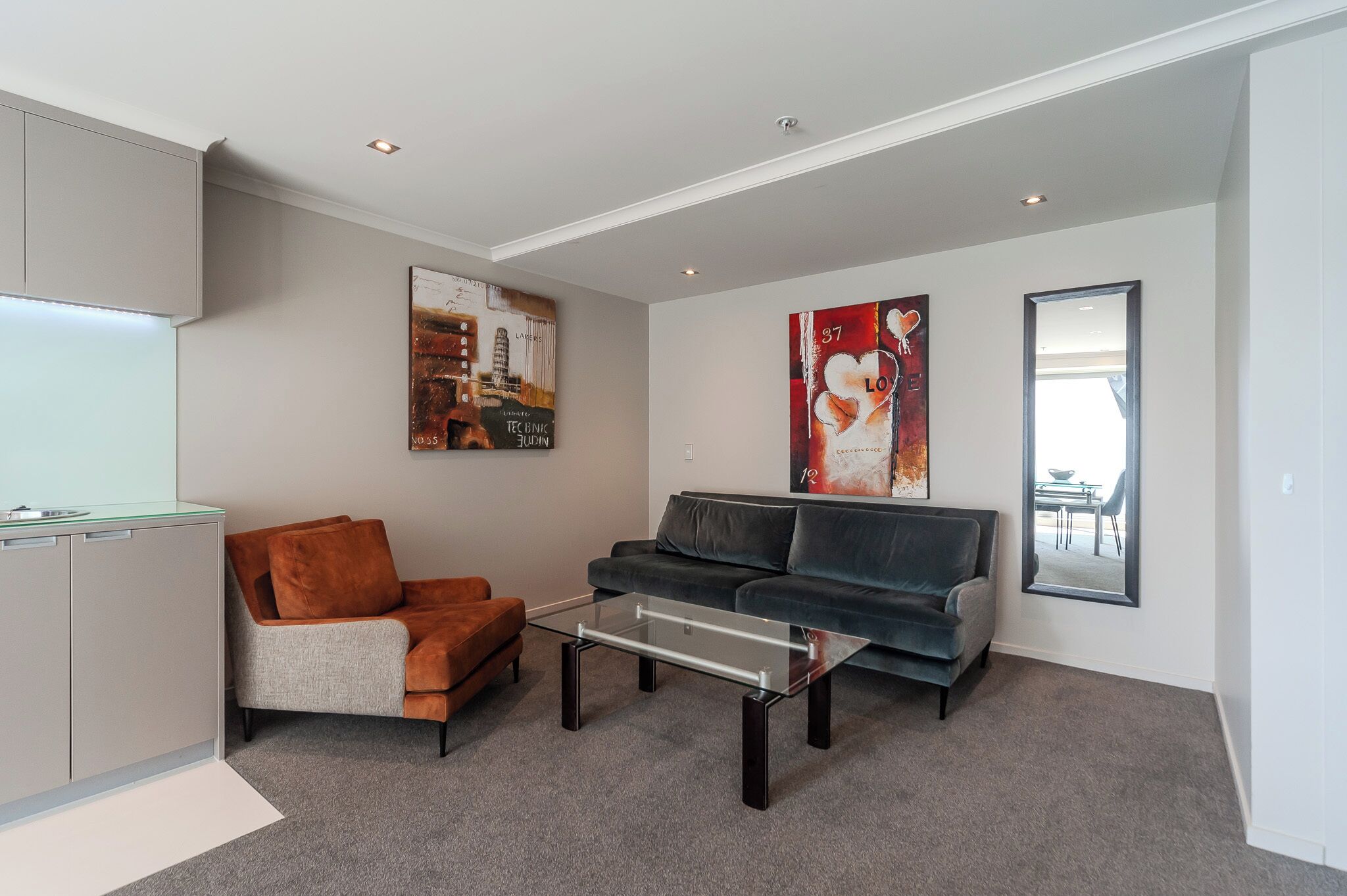A modern living room with a glass coffee table, a grey sofa, and a brown armchair. There are two large paintings on the walls and a large mirror.
