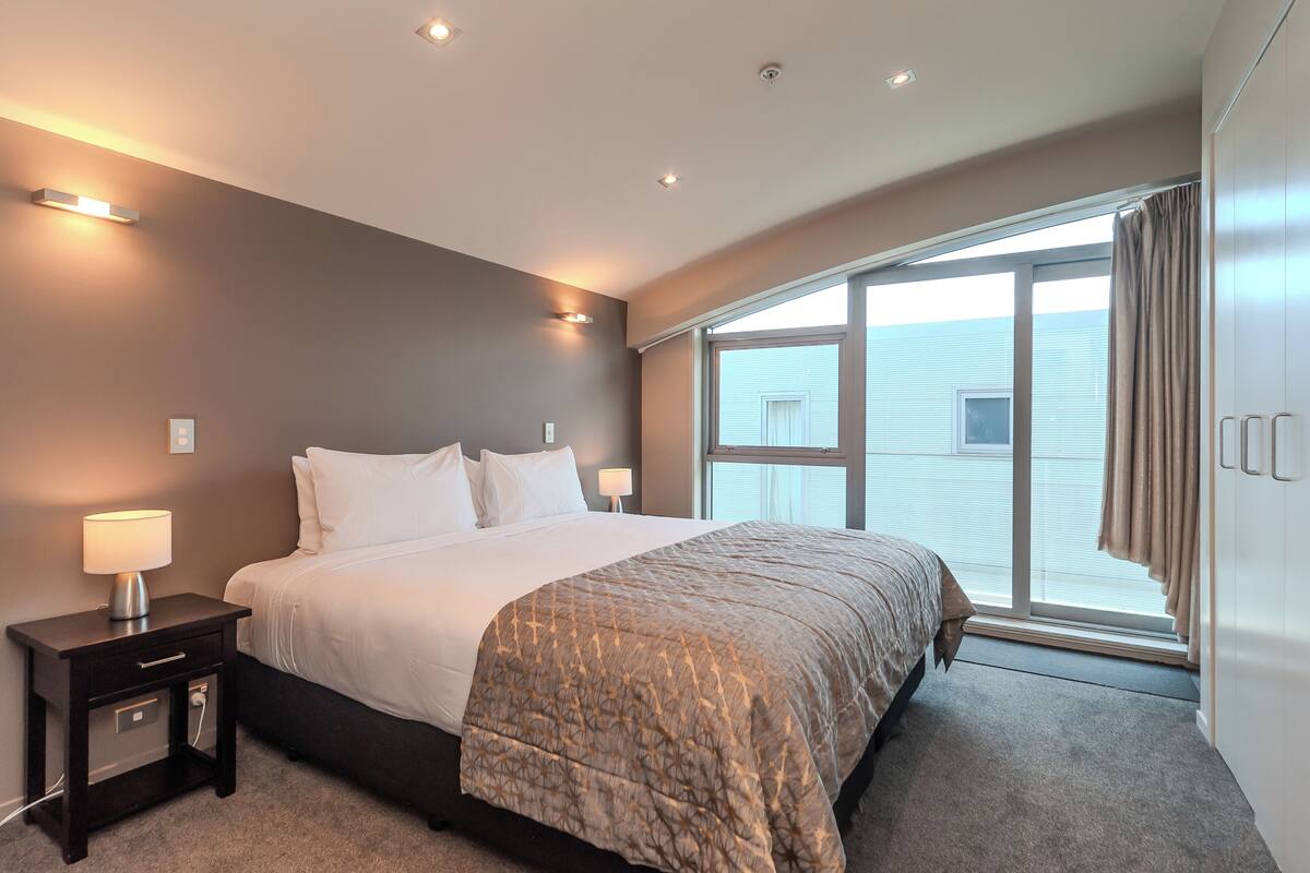 A modern hotel room with a large bed, bedside lamps, a nightstand, and a view of the outside through a large window.