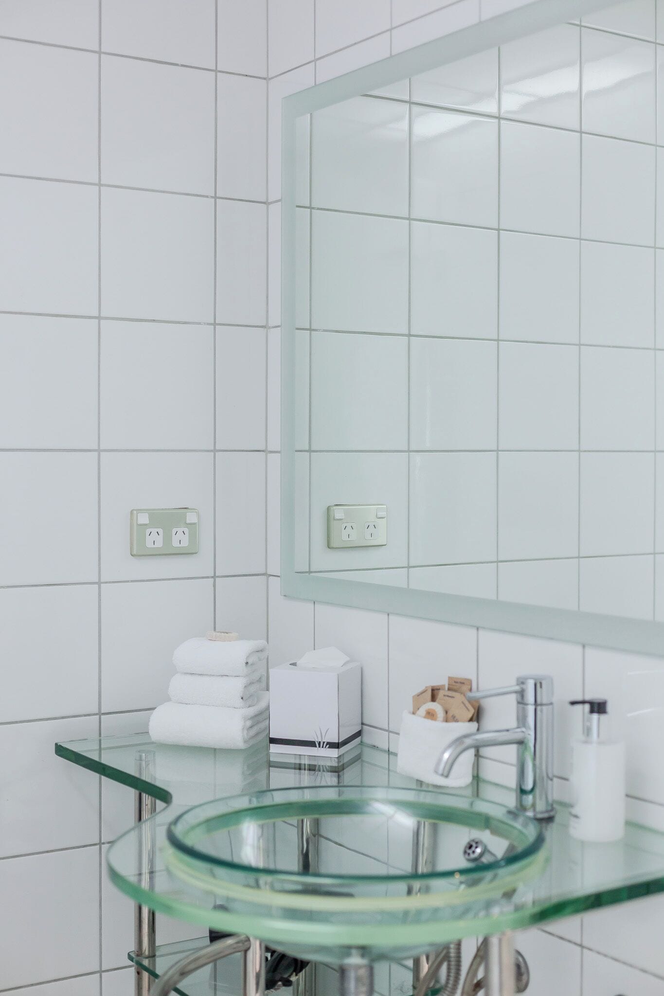 A modern bathroom with white tiled walls, a glass sink, a large mirror, and a towel rack with folded towels.