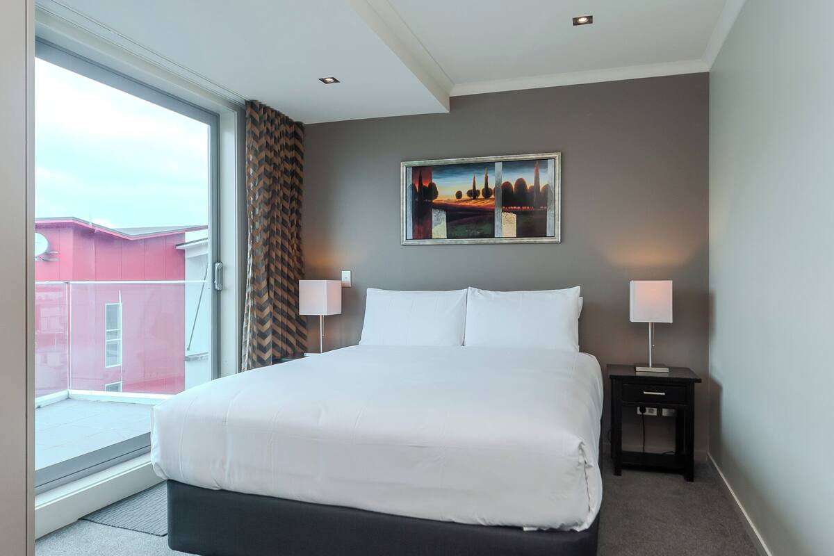 A hotel room with a large bed, two bedside tables with lamps, a painting on the wall, and a view of a red building through a window.