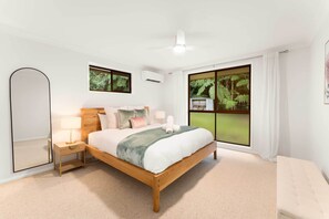 A bedroom with a wooden bed, a nightstand with a lamp, a large window with a view of greenery, and a ceiling fan.