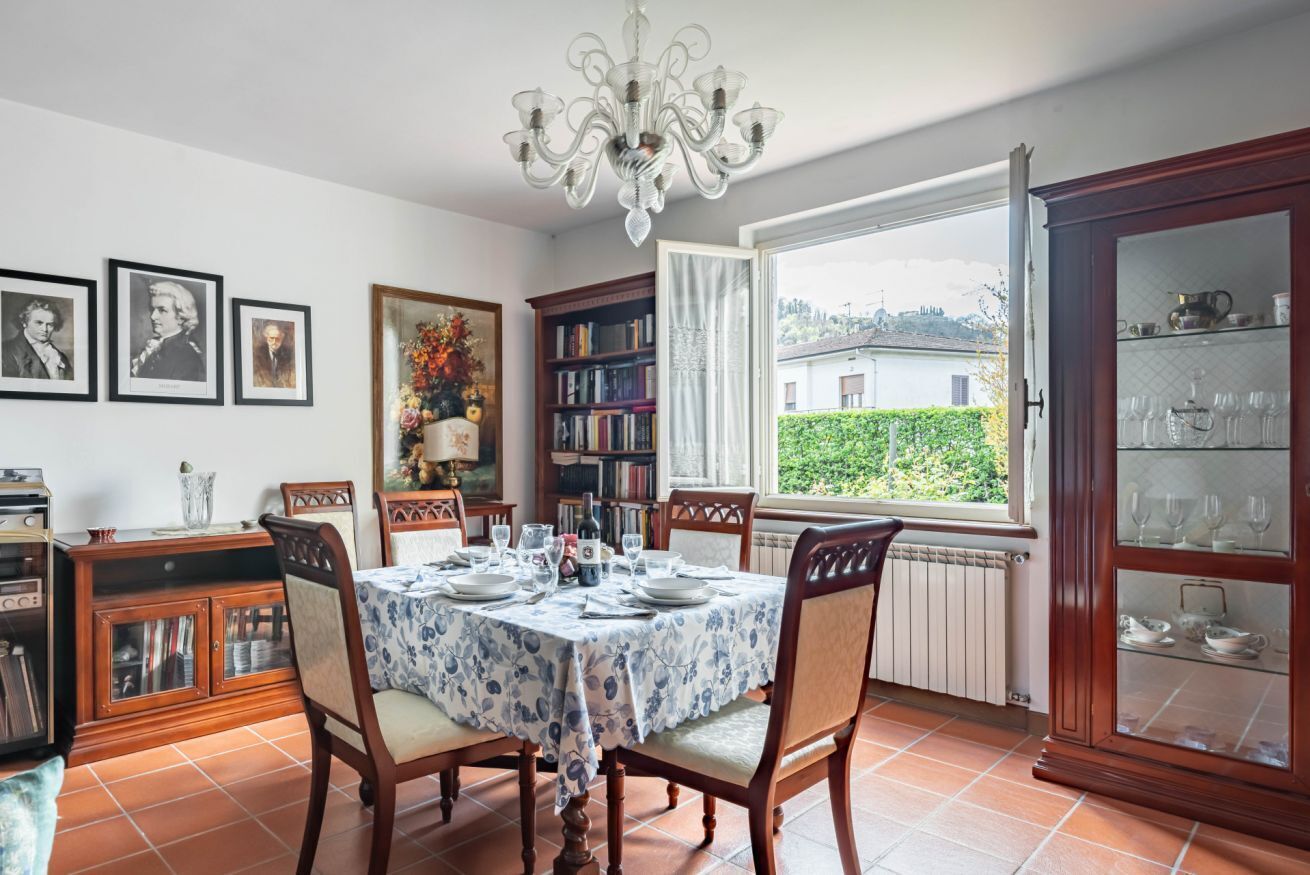 Una sala da pranzo con un tavolo apparecchiato per quattro persone, un lampadario, una libreria e vista su una casa attraverso una finestra.