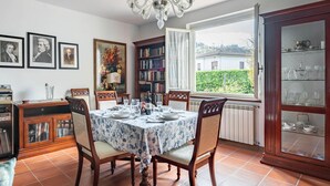 Een eetkamer met een tafel gedekt voor vier personen, een kroonluchter, een boekenkast en uitzicht op een huis door een raam.