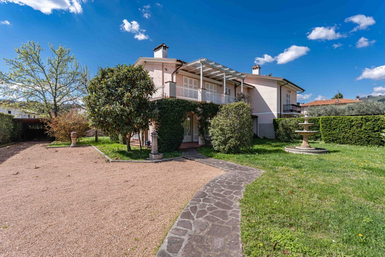 Una casa a due piani con un vialetto in pietra che conduce all'ingresso, immersa nel verde e sotto un cielo sereno.