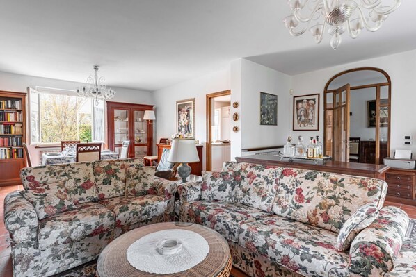 A living room with a floral sofa, a round coffee table, a chandelier, a dining area with a table and chairs, and a kitchen area with a mirror and cabinets.