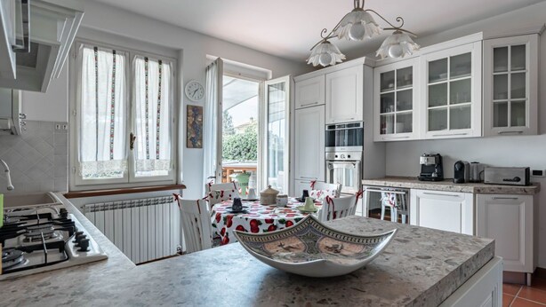 Een keuken met witte kastjes, een gasfornuis, een eettafel met een tafelkleed met dessin en een decoratieve schaal op het aanrecht.