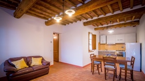 A living room with a brown leather sofa, a dining table with chairs, a kitchen area with cabinets and a refrigerator, and a wooden ceiling.