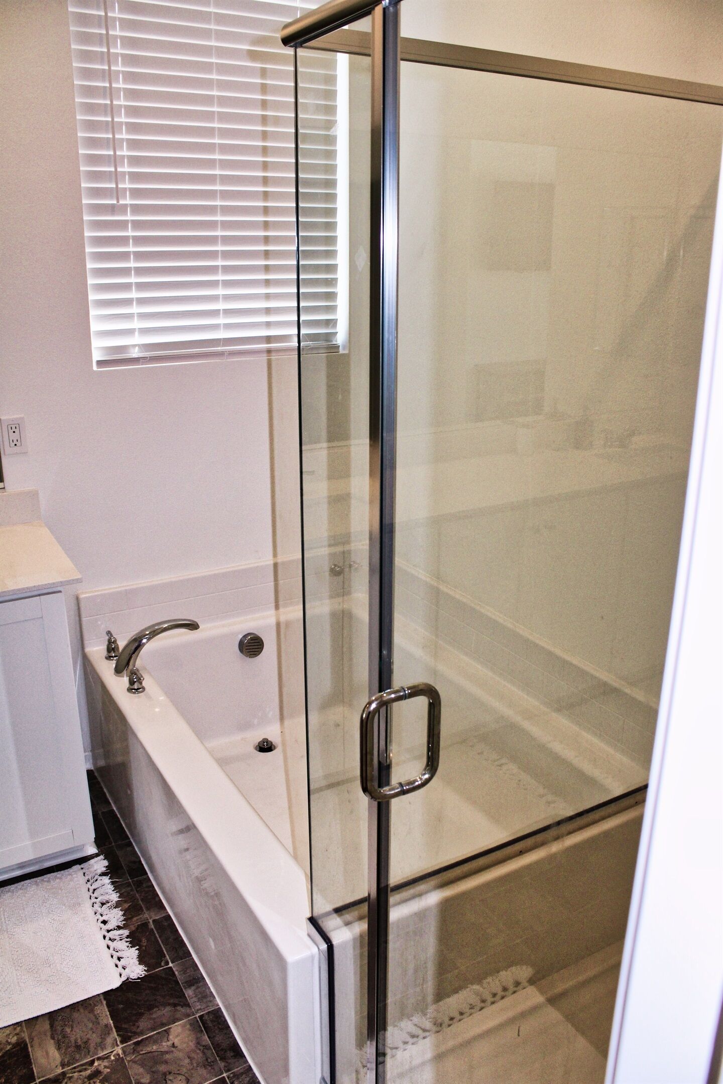 Ein modernes Badezimmer mit einer gläsernen Duschkabine, einer weißen Badewanne und einem Fenster mit Jalousien.