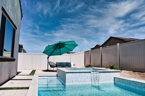 A backyard with a swimming pool, a small hot tub, a patio with chairs, and a large umbrella.