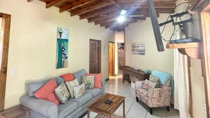 A spacious living room with a high wooden ceiling, a grey sofa with colorful cushions, a patterned armchair, and a wooden coffee table.