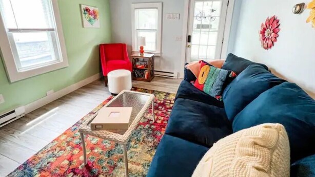 A cozy living room with a blue sofa, a red chair, a small table with a box, and a colorful rug. - Love Shack Private Hot tub  steps to Jet/Town (Port Clinton)