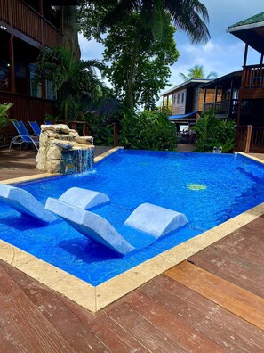Une piscine agrémentée d’une cascade en pierre, entourée d’une terrasse en bois et d’une végétation tropicale.