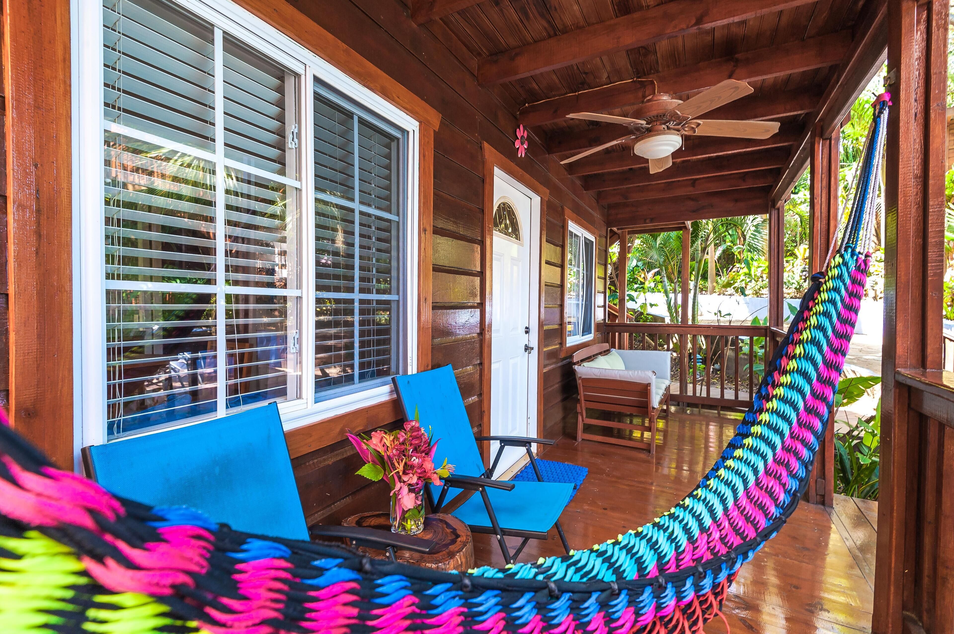 A wooden porch with a hammock, two blue chairs, and a wooden table with a vase of flowers.