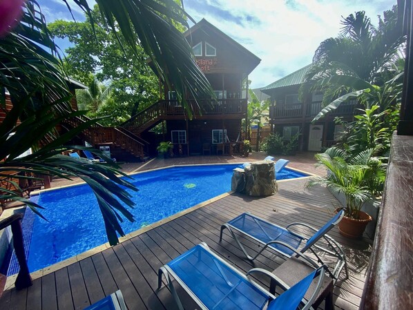 Un espace piscine doté d’une terrasse en bois, de chaises longues et d’un bâtiment sur lequel est apposée une enseigne indiquant « Île de… ».