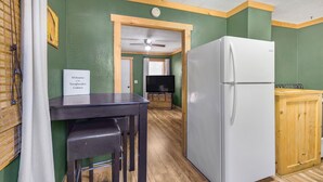A kitchen with a white refrigerator, wooden cabinets, a wooden table with stools, and a television in the background. - Salmon River Cabin #204 (Riggins)