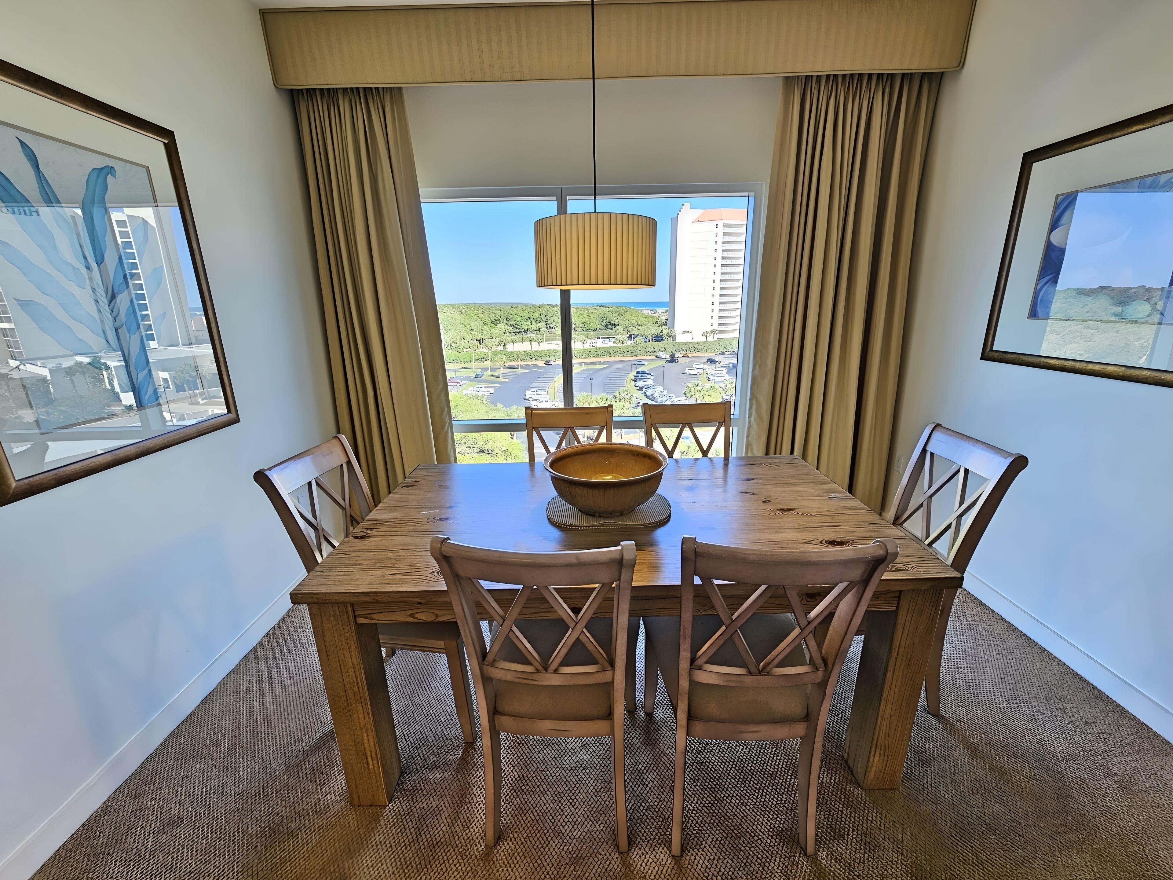 A dining area with a wooden table and chairs, a large woven bowl, and a view of a cityscape through large windows.