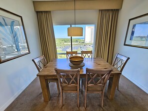 A dining area with a wooden table and chairs, a large woven bowl, and a view of a cityscape through large windows.