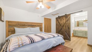 Une chambre avec une tête de lit en bois, un lit recouvert d’une couverture à motifs, un banc en bois, un ventilateur de plafond et une porte coulissante de style grange donnant sur une salle de bain équipée d’un meuble-lavabo et d’un miroir.