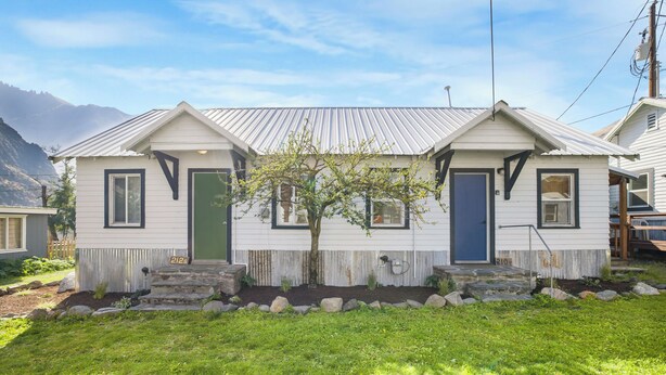 A two-story house with a sloped roof, a small front yard with a tree, and a clear view of the mountains in the background. - Salmon River Cabin #210 (Riggins)