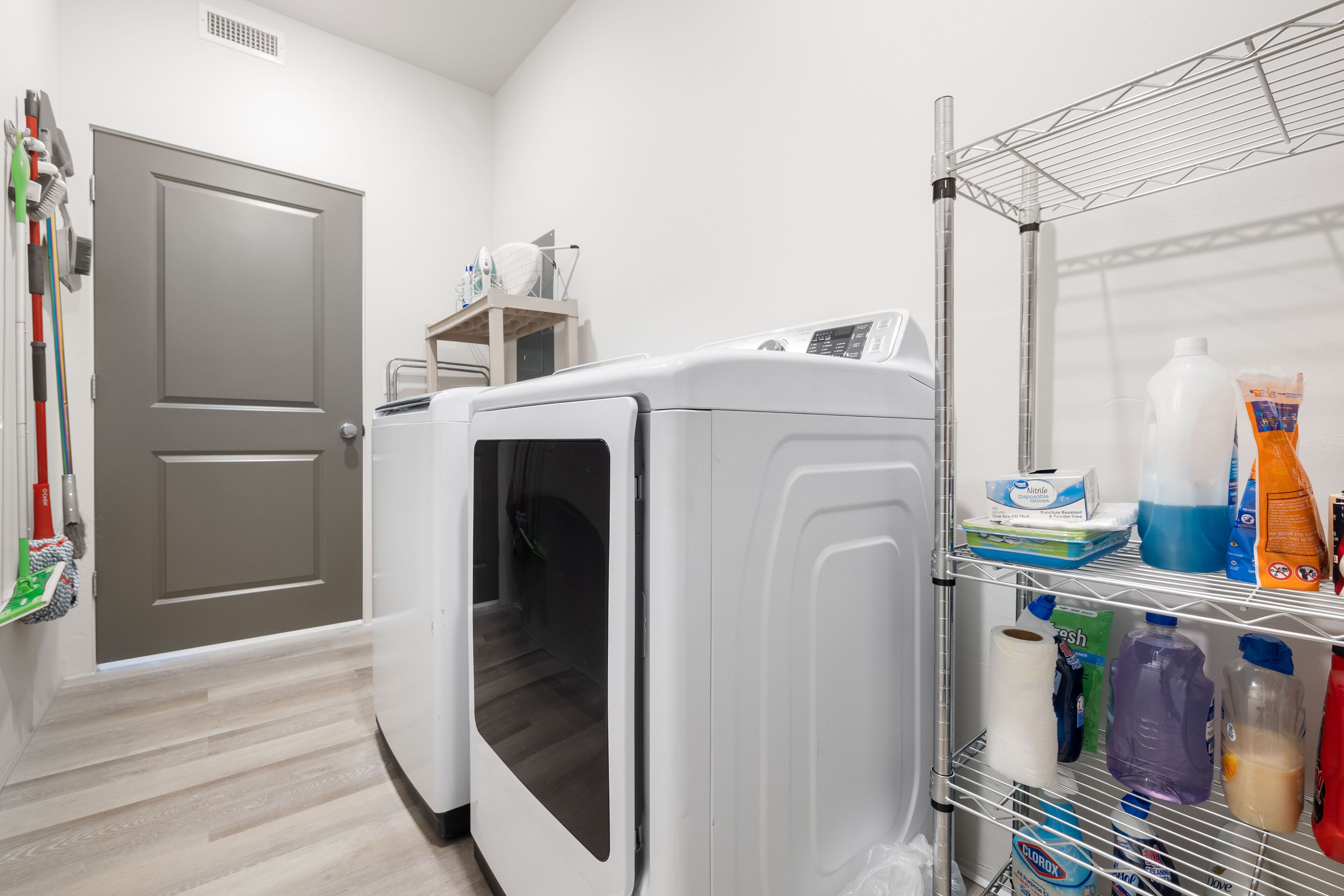 A laundry area with a washing machine and dryer, a shelf with cleaning supplies, and a door with a broom and mop.