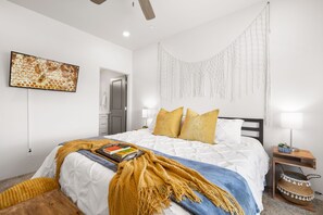 A bedroom with a bed, a wall-mounted TV, a macrame wall hanging, and a bedside table with a lamp and a basket.