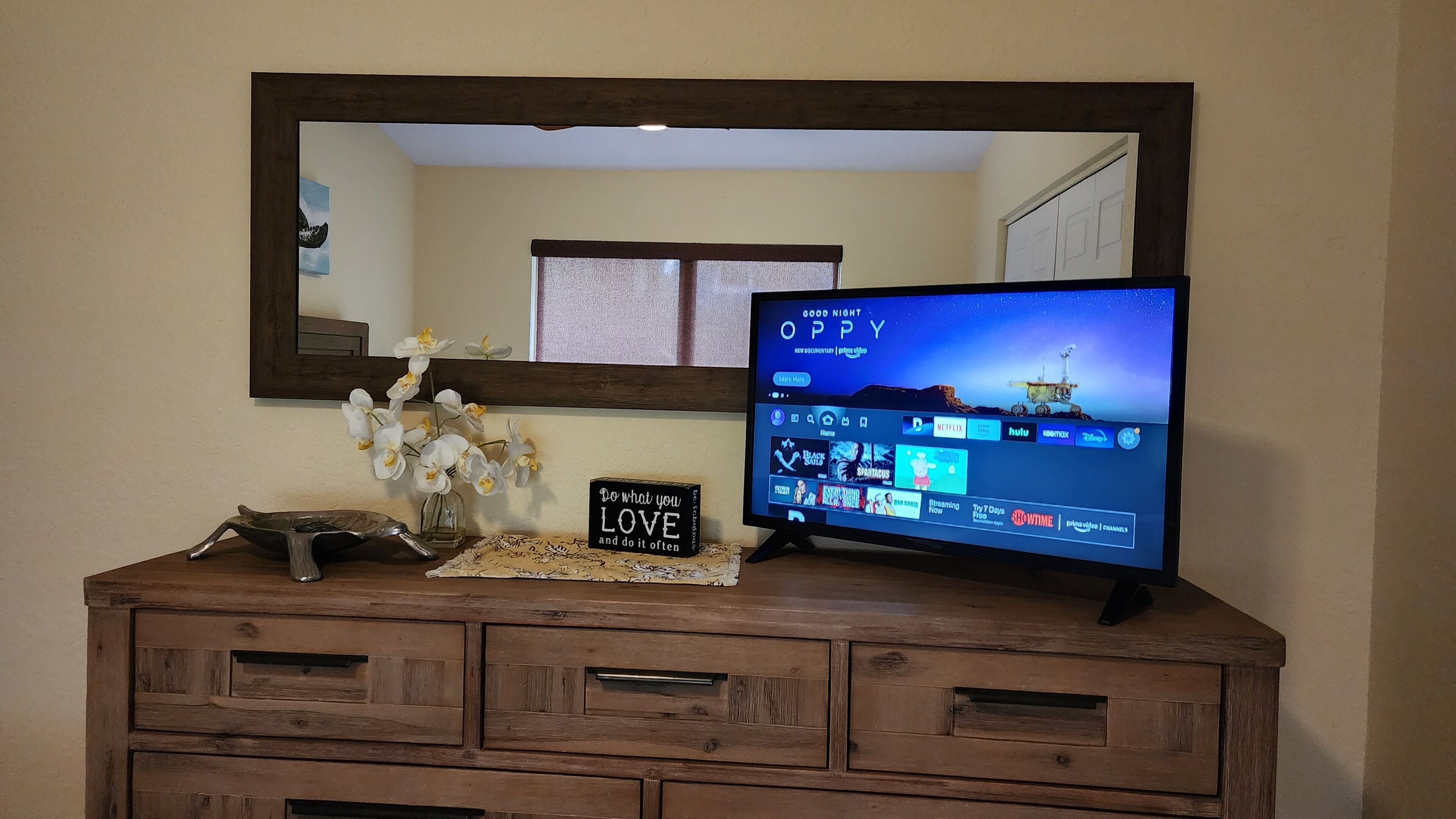 Une commode en bois sur laquelle est posée une télévision à écran plat affichant l’interface d’un service de diffusion en continu, une orchidée décorative et un panneau d’affichage avec un message motivant.