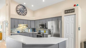 Una cocina moderna con gabinetes grises, una isla grande con taburetes, electrodomésticos de acero inoxidable y un reloj de pared.