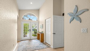 Un pasillo con piso de madera, una puerta blanca, una puerta de vidrio con vistas, una decoración de estrella de mar y una pequeña mesa de madera con una planta.