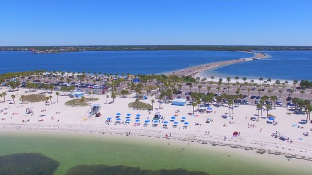 Vista aérea de una playa con zona de estacionamiento, palmeras y agua azul cristalina.