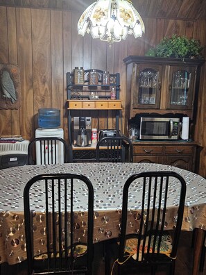 Un coin repas en bois comprenant une table, des chaises, un distributeur d’eau, une machine à café, un four à micro-ondes et un lustre.