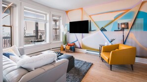 Una sala de estar moderna con un sofá, un reposapiés y un sillón amarillo. Un televisor de pantalla plana está montado en la pared, y hay una mesita con una lámpara.