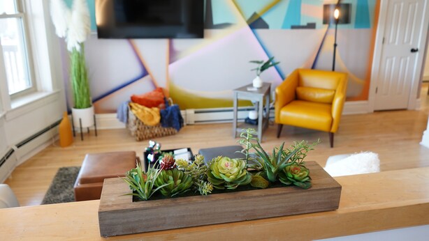 Una sala de estar con una mesa de centro de madera, un sillón amarillo, un televisor de pantalla plana montado en la pared y un diseño abstracto y colorido en la pared.
