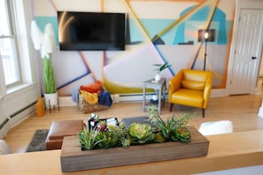 A living room with a wooden coffee table, a yellow armchair, a flat-screen TV mounted on the wall, and a colorful abstract wall design.