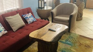 Una sala de estar con un sofá rojo, una mesa de centro de madera y un sillón beige. Hay cuadros enmarcados en la pared y una cocina al fondo.