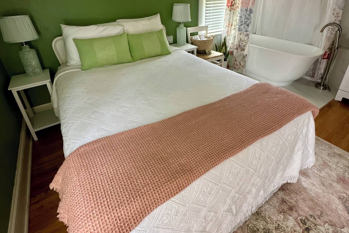 A bedroom with a large bed, a nightstand, a lamp, a window with curtains, and a bathtub.