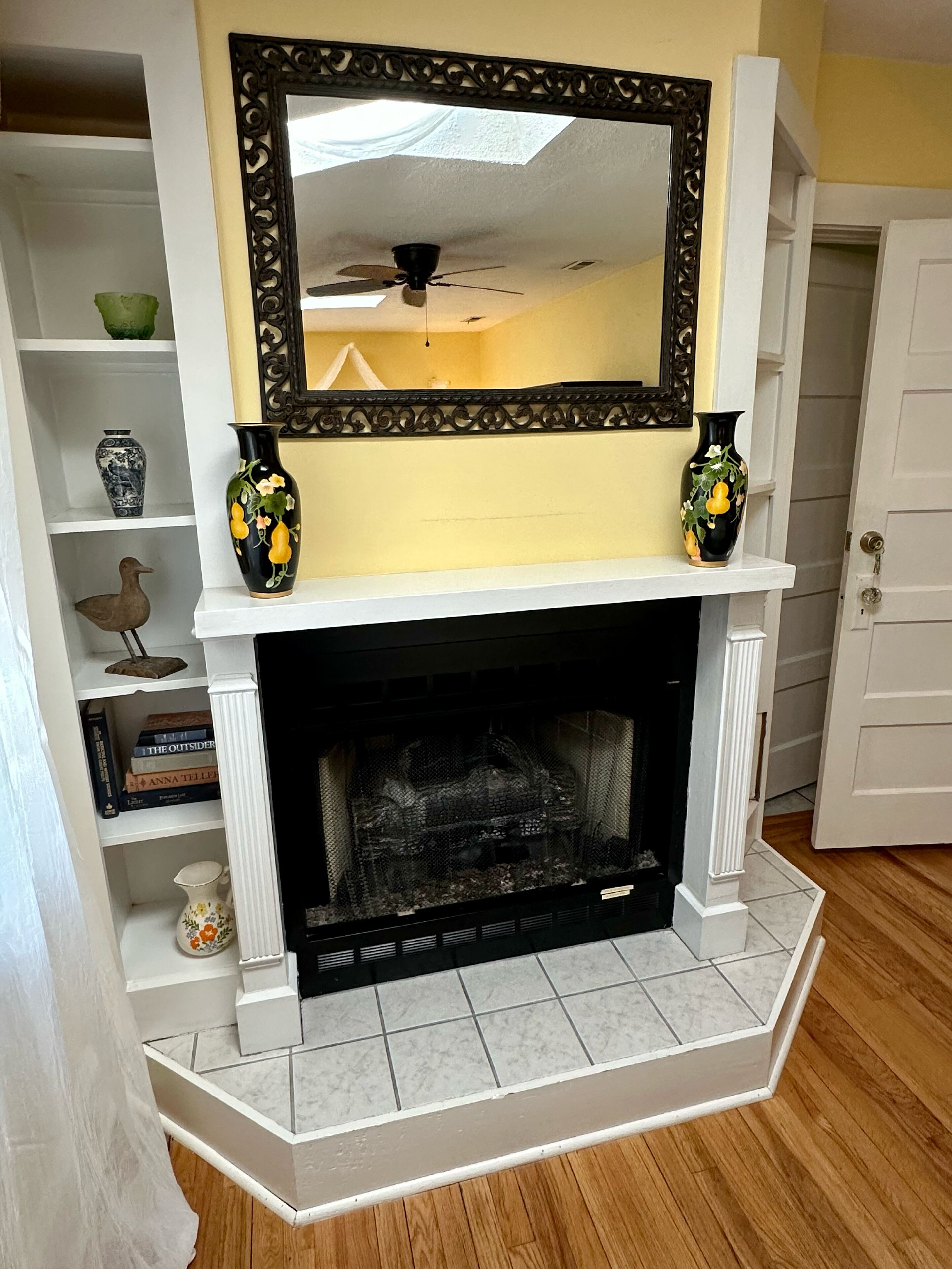 A fireplace with a white mantel, two vases with floral designs, and a bookshelf with various items.