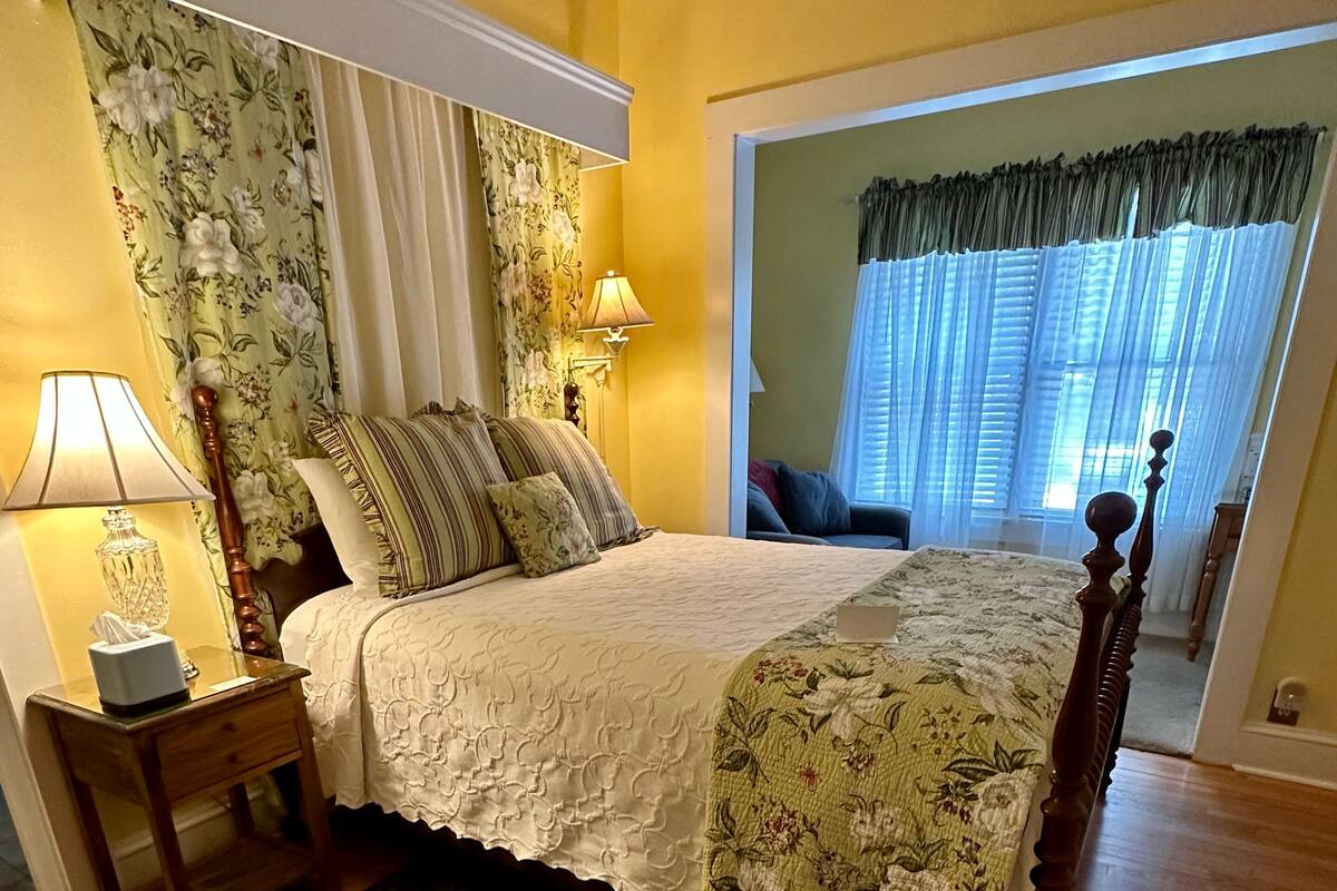 A bedroom with a wooden floor, a bed with floral bedding, a nightstand with a lamp, and a window with sheer curtains.