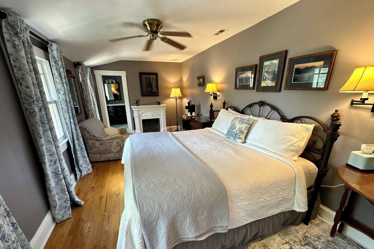 A bedroom with a large bed, a ceiling fan, a fireplace, and a nightstand with a lamp.