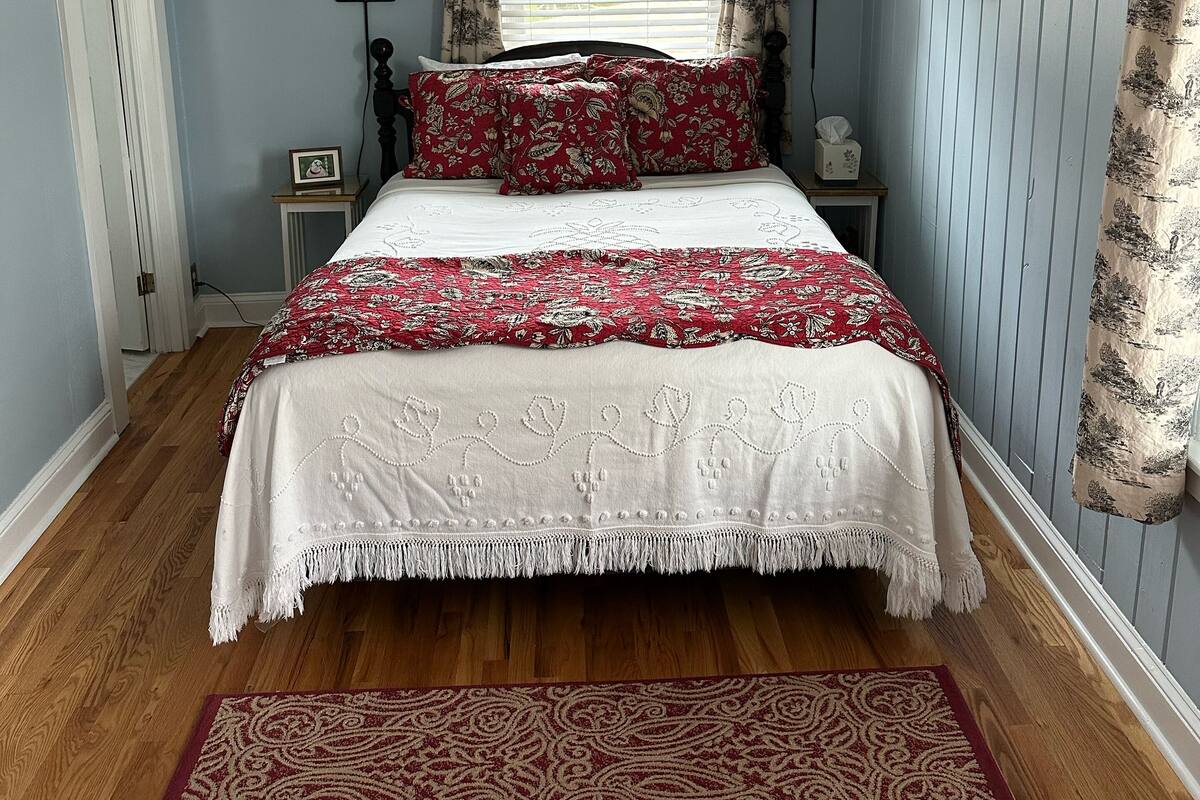 A bedroom with a bed, a ceiling fan, a window with curtains, a bedside table with a lamp, and a framed picture on the wall.
