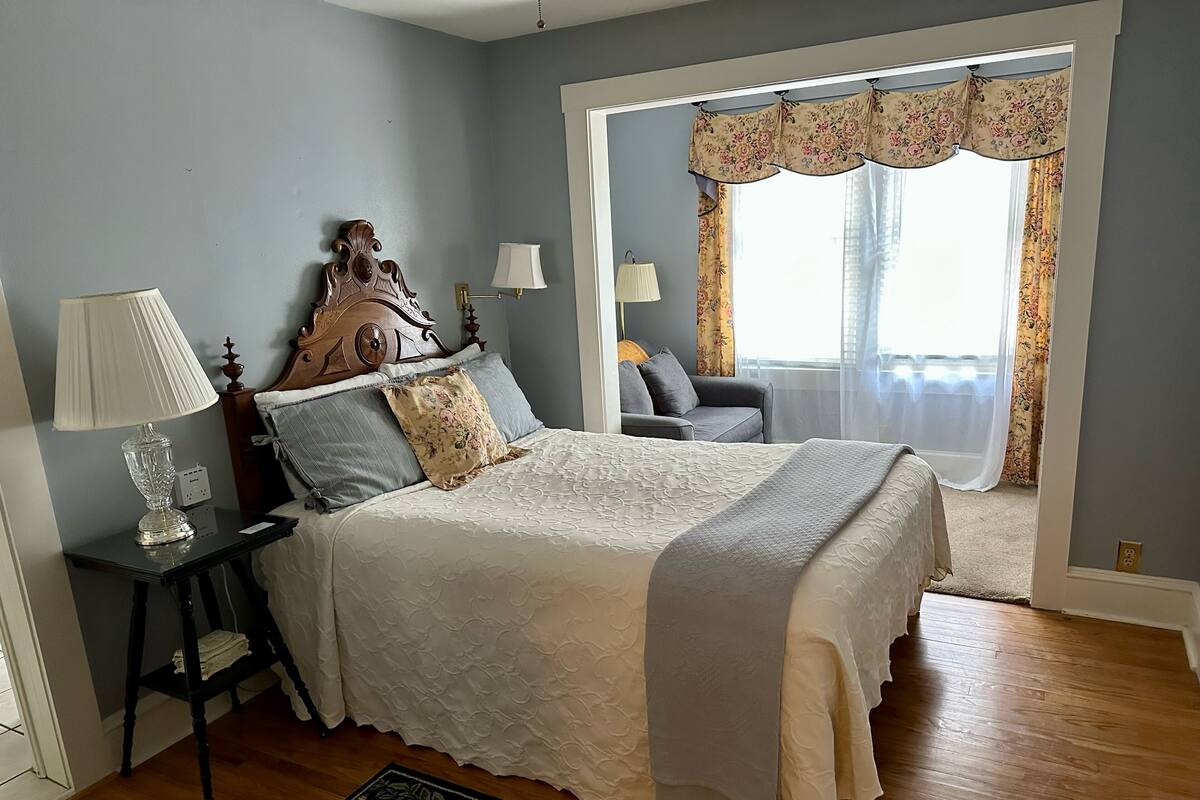 A bedroom with a bed, bedside lamps, a nightstand, a chair, and a window with curtains.