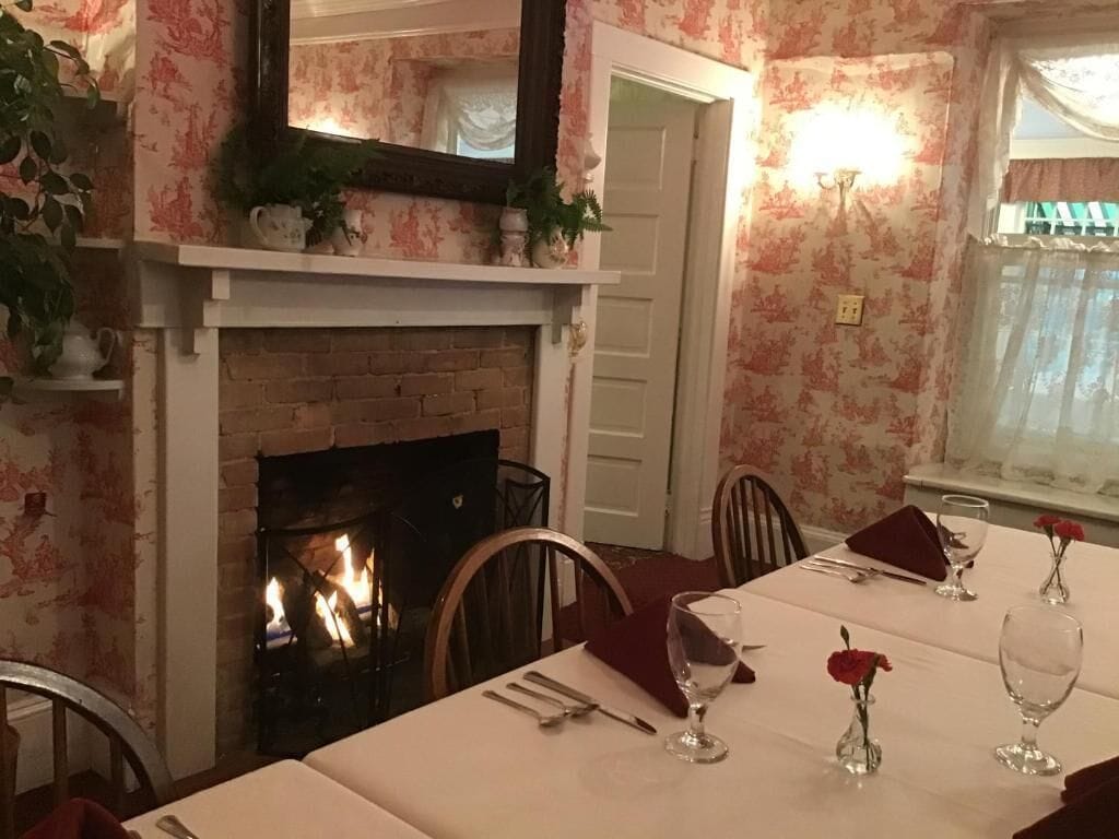 Un comedor con chimenea de ladrillo, papel tapiz floral y una mesa dispuesta para cuatro, con copas de vino y un florero de rosas rojas.