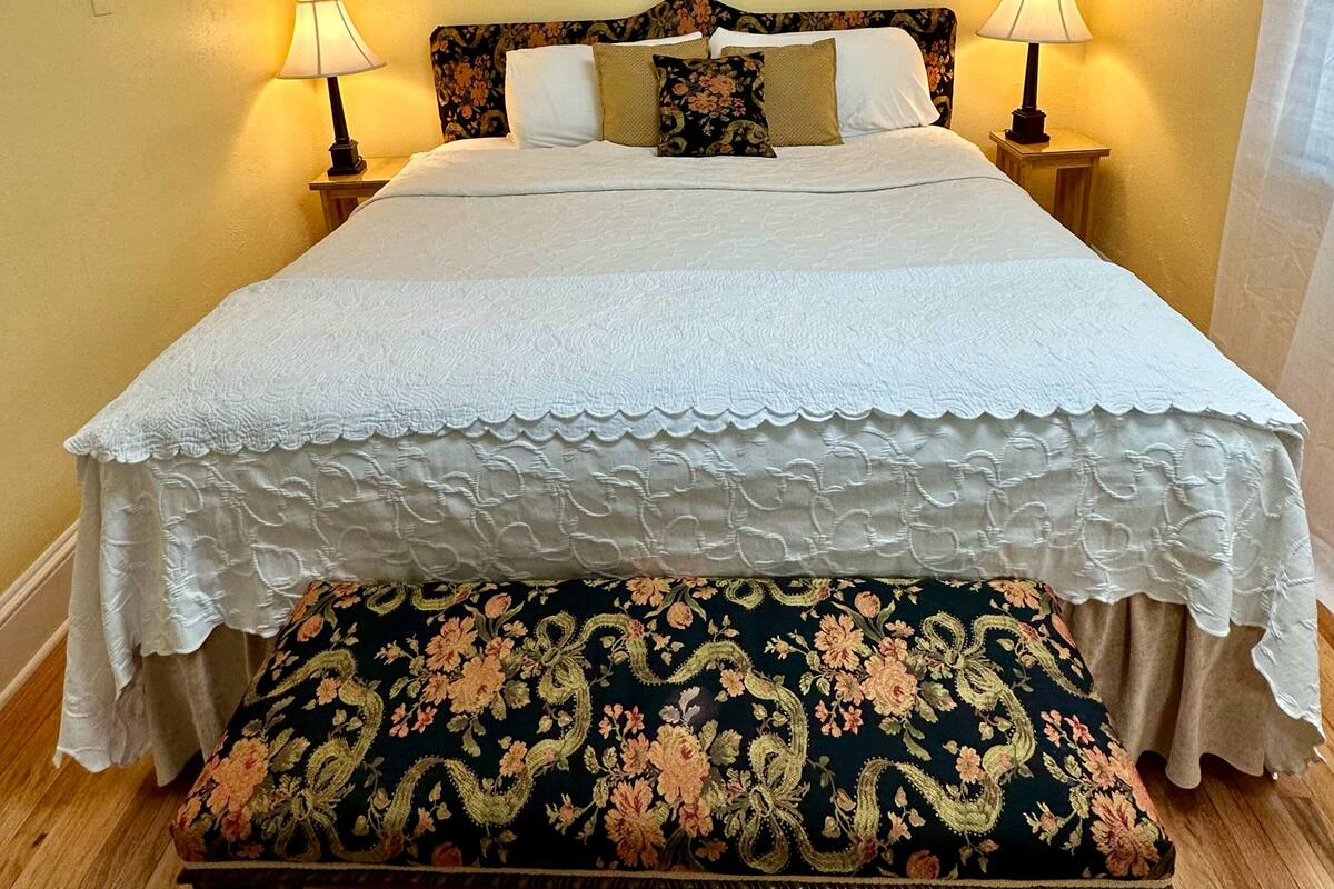A bed with white linens, two bedside lamps, a floral headboard, and a bench with a floral pattern.