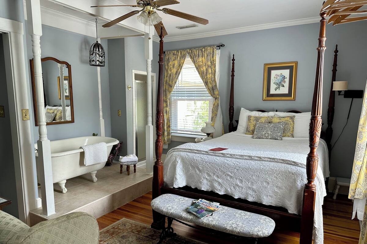 A four-poster bed with a footboard, a sitting bench, a white bathtub, and a ceiling fan.